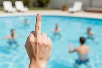 Middle finger hand gesture of rejection against blurred tourists in a swimming pool, showing social isolation and contempt amidst sunny resort leisure, emotional dissonance between collective recreational joy and individual hostility, harsh daylight illum [IBR125446496]