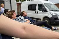 Young travellers resting outside camper vans, road trip fatigue and vanlife leisure culture, communal relaxation during summer vacation journey, nomadic recreation and temporary escapism, reclining friends experiencing collective lethargy and serene repos [IBR125446499]