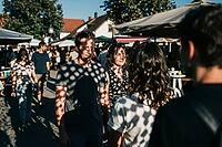 Outdoor market gathering in warm sunlight, visitors chatting on cobblestone pavement under canvas canopies, polka dot shadows filter through perforated shades creating geometric patterns across human faces and casual summer clothing, intense eye contact b [IBR125446503]