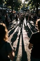 Backlit pedestrian walking alone in crowded promenade, long shadows stretch forward on asphalt pavement, solitary figure gestures while navigating dense urban masses under golden evening light, atmospheric counterlight produces dramatic silhouette contras [IBR125446506]