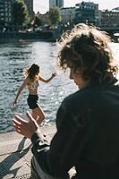 Youthful vitality meets urban waterside escapism, blurred figures engaging in spontaneous gestures while sparkling sunlight reflects off ripples, a dancing woman in light attire balances gracefully along the stone riverbank embankment, shallow depth of fi [IBR125446510]