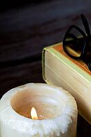 Burning candle providing warm light for a vintage book and reading glasses, symbolizing education, knowledge, and quiet study [IBR125448817]