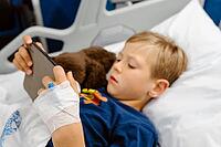Sick boy lying in a hospital bed, playing on his phone with a teddy bear beside him, Portugal [IBR125448829]
