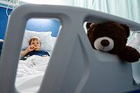 A young boy resting and eating in a hospital bed while holding a teddy bear [IBR125448843]