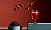 Glass of iced red drink sits on a white surface. Behind it, a branch with dried hops hangs. Cluster of dark red grapes rests nearby, AI generated [IBR125448877]