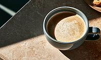 Sunlight highlights coffee cup on textured stone surface. Smooth froth sits atop dark coffee liquid, AI generated [IBR125448880]