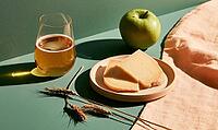 Glass of beer beside green apple and cheese slices on wooden plate. Warm sunlight casts soft shadows on green and pink surface, AI generated [IBR125448886]