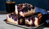 Three square blueberry cheesecake pieces on metal tray. Berry sauce drips over golden crust. Dark coffee mug sits behind, AI generated [IBR125448893]