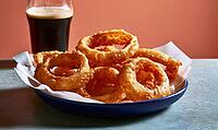 Crispy fried onion rings sit in blue dish. Glass of dark beer sits beside. Food rests on white paper. Background is warm orange wall, AI generated [IBR125448897]