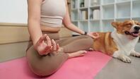 Close-up of woman sitting in meditation posture with mudra hand gesture on pink exercise mat while her dog rests next to her [IBR125462052]