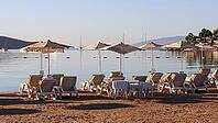Beach chairs and umbrellas line a sandy shore, overlooking a calm sea with boats and distant hills under a clear sky. [IBR125462056]