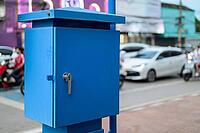 A vibrant blue metal electric or supply box mounted on a pole outdoors, set against a blurred background of urban traffic and street side shops [IBR125462058]