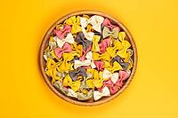 Farfalle pasta of various colors filling a wooden bowl, presented as dry ingredients on a vibrant yellow background and offering a cheerful culinary theme [IBR125462086]