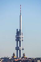Prague, Czech Republic - February 15, 2019: The Zizkov Television Tower rises above the rooftops of the Zizkov district against a clear blue sky, with church towers and residential buildings visible at the base [IBR125462089]