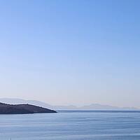 A tranquil blue seascape with a dark coastline in the foreground, and hazy distant mountains fading into a clear, gradient sky. [IBR125462096]