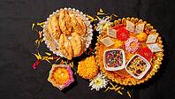 A top-down view of traditional Indian sweets on silver and gold platters, a burning clay diya, marigold flowers, and bowls of sprinkles on a black background [IBR125462097]