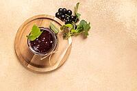 Black currant jam, prepared at home, filling a glass jar on a wooden serving board, featuring fresh berries and green leaves, representing sweet fruit preservation and delicious dessert spread [IBR125462103]