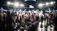 Political campaign rally inside a crowded sports arena with broadcast camera equipment, television journalists and a clapping politician in the center, media spectacle and public relations staging within modern democratic election processes, large televis [IBR125472246]