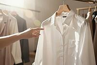 Tactile textile appreciation and sustainable capsule wardrobe curation define mindful consumption in contemporary fashion retail, where natural morning sunlight illuminates a pristine white cotton blouse on a smooth wooden hanger, an anonymous outstretche [IBR125472250]