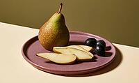 Single pear and sliced fruit on purple plate. Three black grapes beside pear. Warm lighting highlights textures, AI generated [IBR125472253]