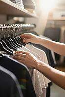 Deliberate tactile selection of monochrome cotton garments on a metal clothes rack represents conscious consumerism and aesthetic wardrobe curation, anonymous hands gently sift through casual apparel suspended on uniform black hangers under warm boutique  [IBR125472259]