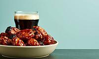 Bowl of glazed chicken wings with beer glass behind. Wings sit on white plate against teal background. Food photo with warm lighting, AI generated [IBR125472268]