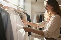 Conscious retail shopping in boutique fashion showroom, elegant woman inspecting white textile shirt on garment rack, slow fashion apparel curation, tactile evaluation of finely woven natural fibres reflects a deliberate shift away from fast fashion towar [IBR125472271]