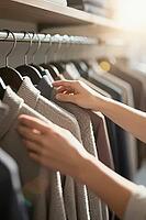 Tactile browsing through knitted sweaters on wooden hangers in a curated capsule wardrobe, soft merino wool garments arranged by neutral earthy tones, gentle fingers touching ribbed cuffs and woven textures symbolize mindful consumerism contrasting dispos [IBR125472275]