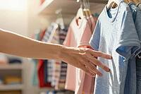 Tactile evaluation of cotton shirts on wooden hangers defines sensory retail consumption, an outstretched arm reaches for soft blue apparel fabrics, selective consumer touch bridges emotional desire with material durability, daylight illuminates delicate  [IBR125472277]