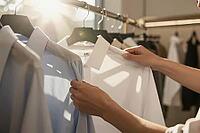 Mindful wardrobe curation, tactile fabric inspection of crisp cotton shirts on metal clothes rack reflects conscious lifestyle and personal dressing, delicate hands touch pale blue and clean white woven textiles suspended in warm natural morning sunlight, [IBR125472281]