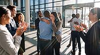Emotional business team celebration in modern glass corporate lobby, diverse colleagues applauding and embracing tearful woman after promotion success, authentic human connection softening rigid corporate architecture, sunbeams flooding through high windo [IBR125472282]