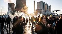Climate justice activism confronting fossil fuel infrastructure, riot police lines facing shouting youth protesters amid drifting smoke and blinding lens flare, massive cooling towers and industrial smokestacks towering above turbulent civil disobedience  [IBR125472293]