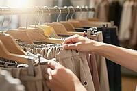 Hands delicately examining woven linen garments on timber coat hangers represent mindful wardrobe choices in contemporary boutique retail environments, sunlight glints across a metallic clothing rail illuminating neutral tones of curated skirts and lightw [IBR125472297]