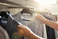 Hands examining light blue pajama shirts on clothing rails in a fashion boutique highlight textile quality, tactile fabric evaluation, retail consumer behavior, intimate apparel selection, wardrobe curation, soft sunlight lens flare illuminating fabric we [IBR125472298]