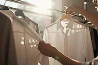 Woven cotton shirts hang on a polished metal garment rack in bright sunlight as hands gently touch soft textile fabrics during clothing selection, tactile sensory examination of high quality tailored garments underscores mindful consumer choices and slow  [IBR125472299]