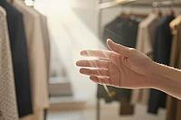 Tactile evaluation of sustainable garments in a minimalist boutique, an outstretched hand catching warm daylight between curated clothing racks, somatic perception meets deliberate retail curation within contemporary slow fashion aesthetics, natural illum [IBR125472300]