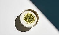 Top-down view of a white bowl with creamy yogurt. Yogurt topped with crushed green pistachios. Bowl placed on white and dark blue surface, AI generated [IBR125472301]