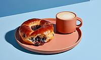 Golden-brown blueberry-filled bagel on terracotta plate. Next to cup of frothy coffee. Set against clean blue background, AI generated [IBR125472306]