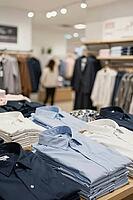 Folded cotton shirts on retail display tables showcase professional menswear and textile merchandising within a modern department store, neat stacks of blue and white button down garments illustrate consumer society dynamics, soft ambient interior lightin [IBR125472311]