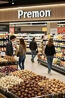 Bulk food market display with nuts and dried snacks represents modern retail nutrition alongside browsing customers in a contemporary grocery hall, foreground arrangements of edible seeds, legumes, dragees, glazed hazelnuts, chocolate confectionery, roast [IBR125472312]