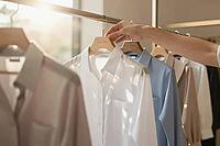 Curated sustainable wardrobe selection, a minimalist white cotton button down shirt hanging neatly on a wooden clothes rail in warm morning light, an anonymous hand thoughtfully browsing essential apparel pieces, elegant pastel textile fabrics representin [IBR125472313]