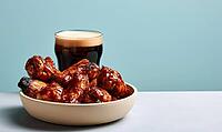 Shiny glazed chicken wings in white bowl. Dark beer with head beside food. Light blue background behind setup, AI generated [IBR125472315]