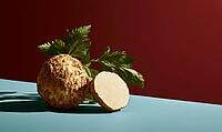 Celeriac root with fresh green leaves on blue surface. One half sliced, showing pale interior. Deep red background contrasts with cool tones, AI generated [IBR125472333]