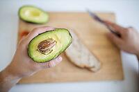 Male hand holding half avocado over blurry wooden cutting board with bread and tomatoes. Man at white table preparing avocado toast. Healthy meal preparation [IBR125492330]