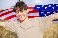 Happy and cheerful teenage boy with arms spread wide waving American flag in a field outside the city. [IBR125492332]