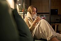 Female seated on sofa wrapped in thick scarf holding a glass of water and smartphone, viewed in warm indoor lighting with a relaxed front angle and visible living room elements [IBR125492347]