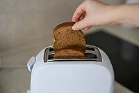 Handhalten Single Brown Bread Slice oben Weiß Zwei Slice Toaster auf Küchenarbeitsplatte in der Nähe von Black Electric Kochplatten im Innenbereich Schließen Sie nach oben Angehobene Slice bereit für Einlegearbeiten [IBR125492365]
