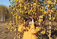 Young woman in yellow hood with binoculars watching birds in autumn forest bird watching, zoology, ecology research, observing animals ornithology [IBR125492371]