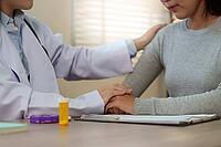 A healthcare professional in a white coat places a reassuring hand on the shoulder of a sad young Asian woman, offering support and empathy during a clinic visit [IBR125492374]