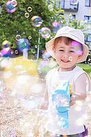 A cheerful toddler boy wearing a bucket hat laughs while shooting soap bubbles from a blue toy gun on a bright, sunny day outdoors [IBR125492375]
